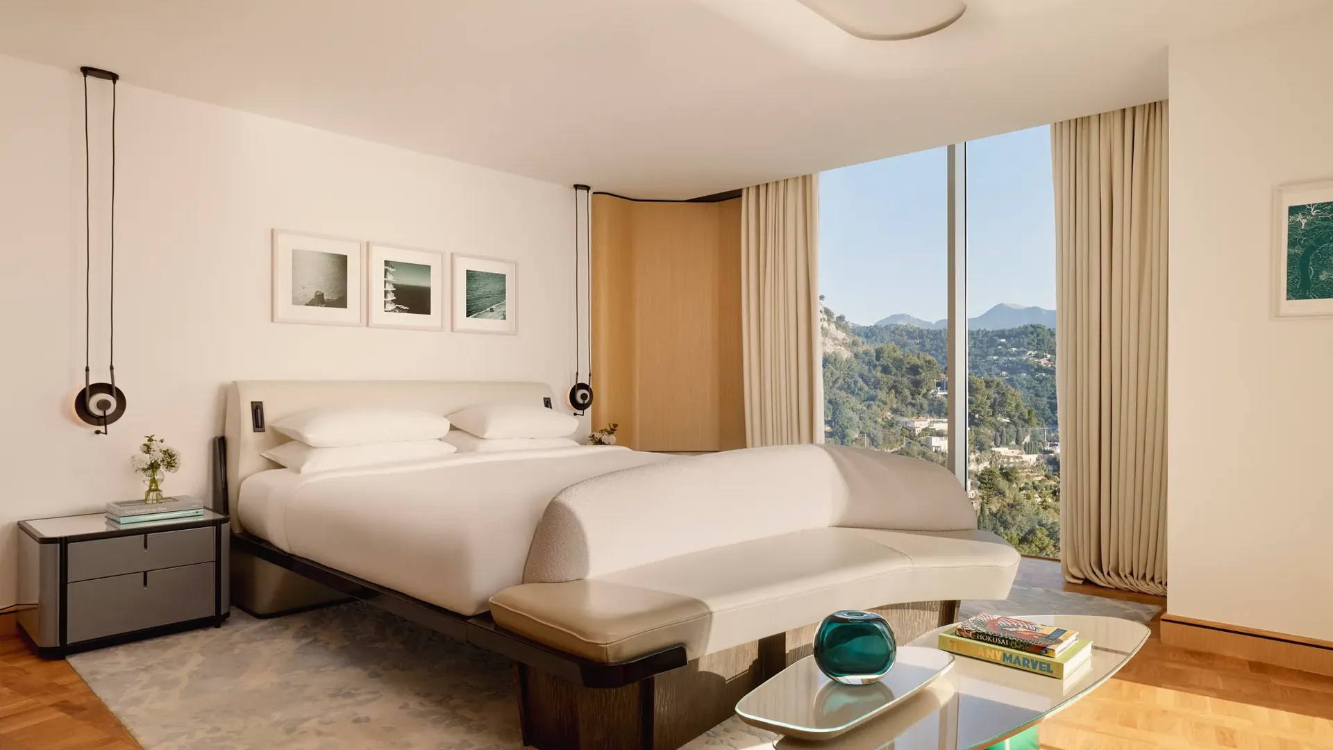 Hotel bedroom with large bed, curved bench, and glass wall offering views of forested hills and mountains, styled in modern neutral tones.