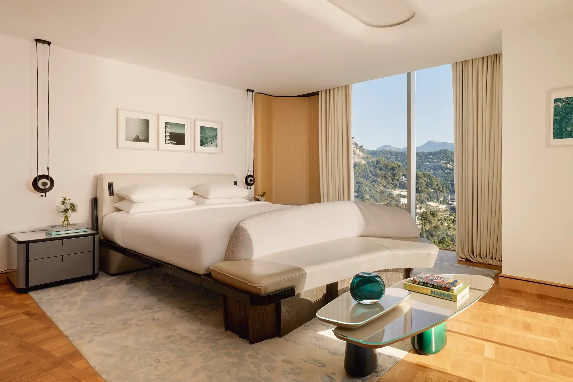 Hotel bedroom with large bed, curved bench, and glass wall offering views of forested hills and mountains, styled in modern neutral tones.