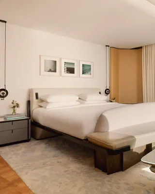 Hotel bedroom with large bed, curved bench, and glass wall offering views of forested hills and mountains, styled in modern neutral tones.