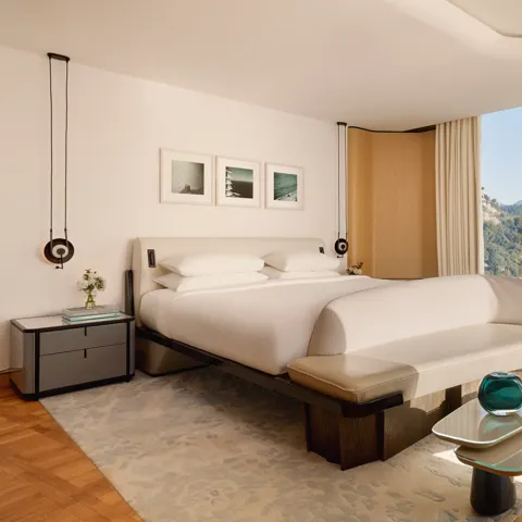 Hotel bedroom with large bed, curved bench, and glass wall offering views of forested hills and mountains, styled in modern neutral tones.
