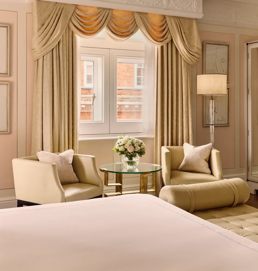Seating nook with two cream armchairs, round glass table, and draped window beside the bed.