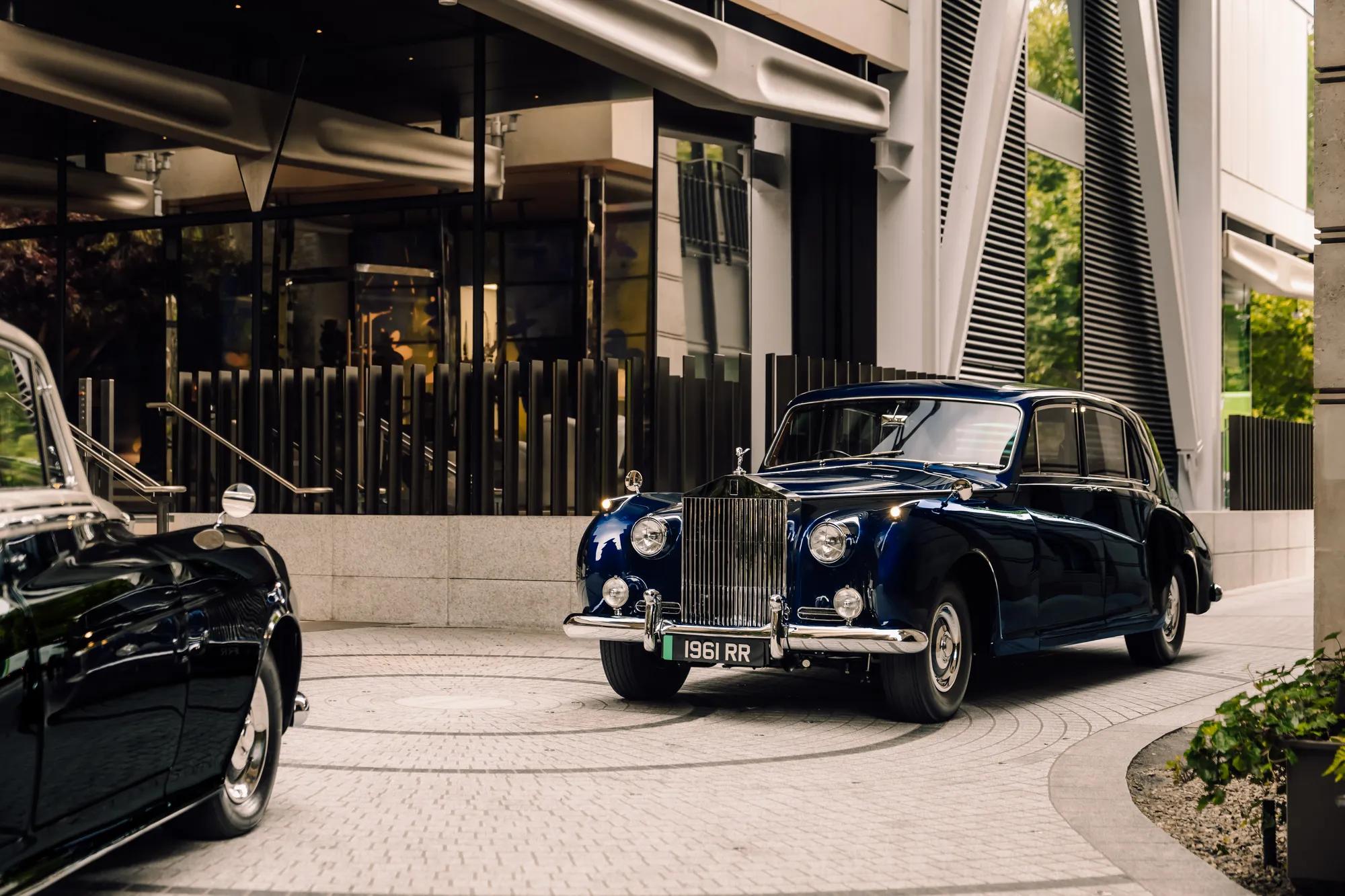A vintage navy blue Rolls-Royce parked at a modern hotel entrance with reflective glass and architectural metal detailing.