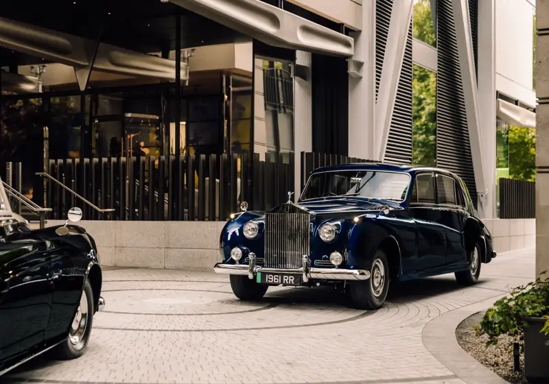 A vintage navy blue Rolls-Royce parked at a modern hotel entrance with reflective glass and architectural metal detailing.
