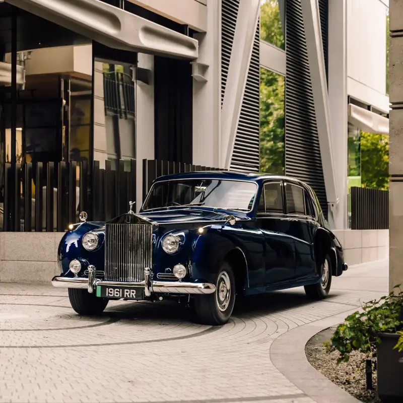 A vintage navy blue Rolls-Royce parked at a modern hotel entrance with reflective glass and architectural metal detailing.