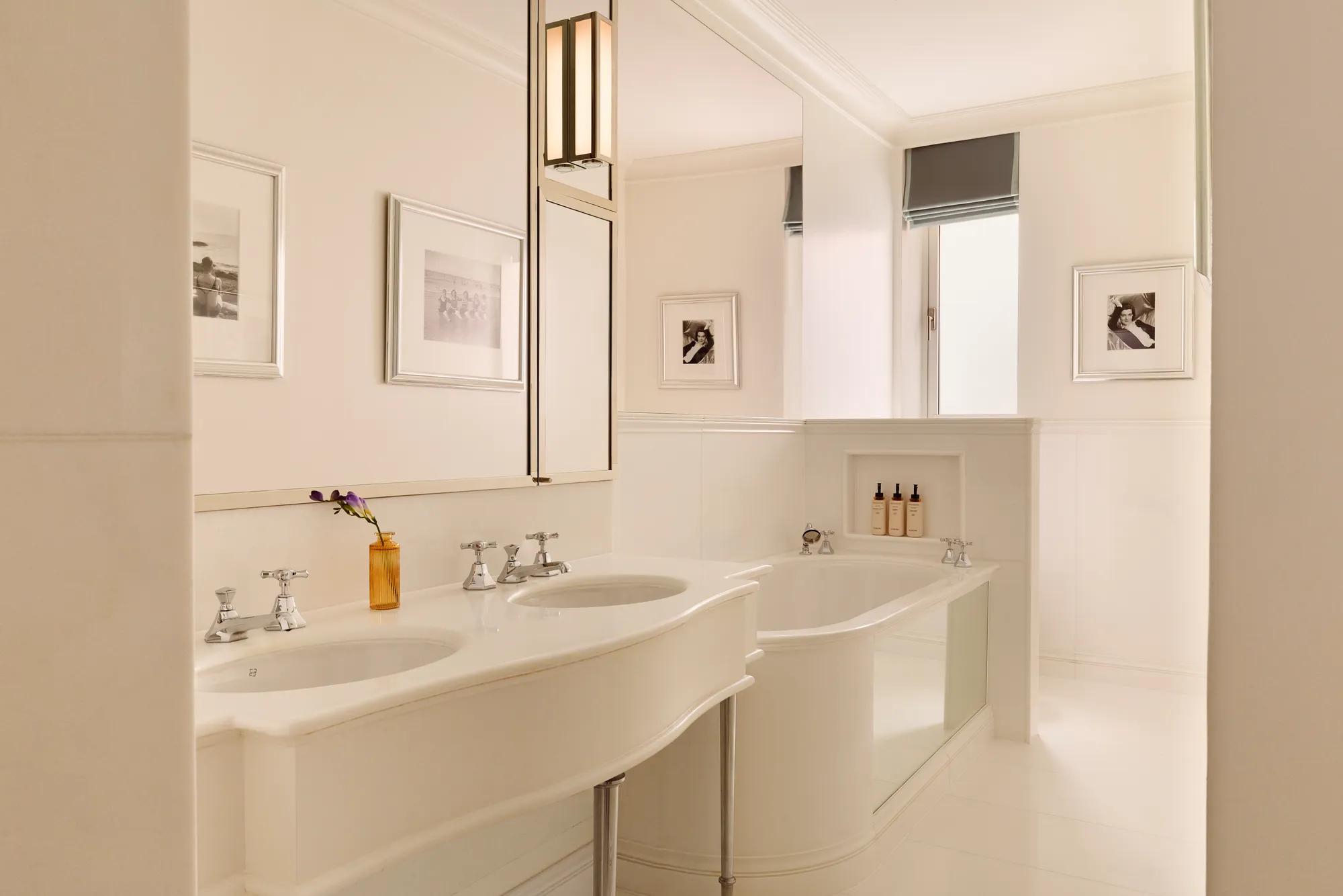 Bright white bathroom with double sinks, framed photos, and a deep tub with bath products set into a wall niche.