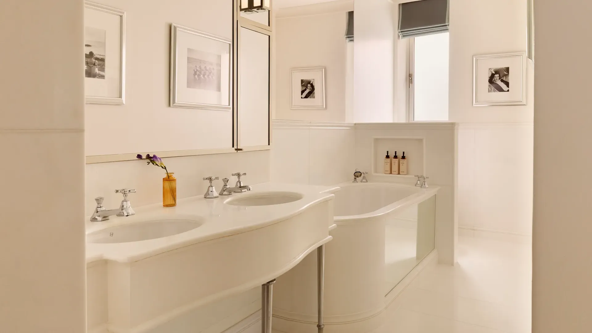 Bright white bathroom with double sinks, framed photos, and a deep tub with bath products set into a wall niche.