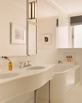 Bright white bathroom with double sinks, framed photos, and a deep tub with bath products set into a wall niche.