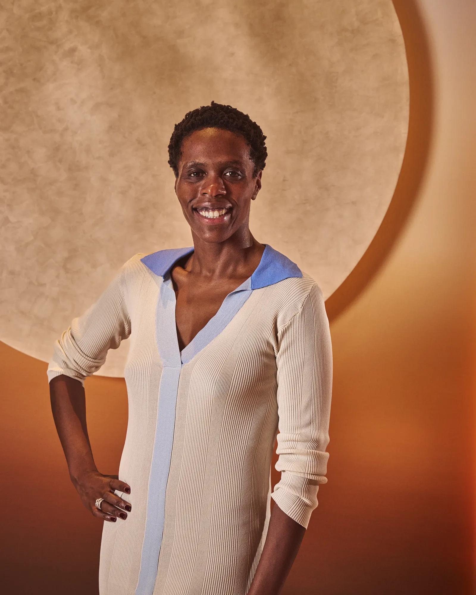 Smiling woman in a cream dress with blue trim stands against a warm-toned background with a circular feature