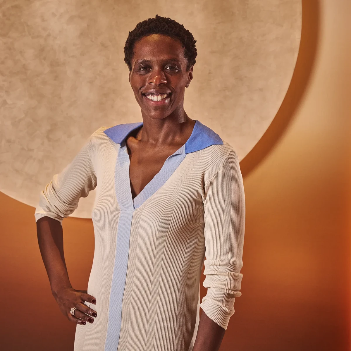 Smiling woman in a cream dress with blue trim stands against a warm-toned background with a circular feature
