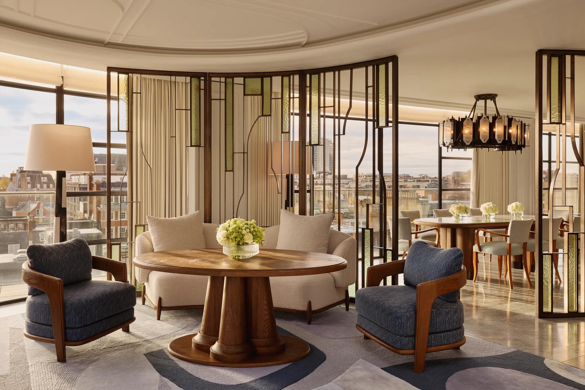 Bright open-plan living and dining area with large curved windows, featuring cream and blue armchairs around a round wooden table, decorative glass partitions, and a marble dining table beneath a statement chandelier.