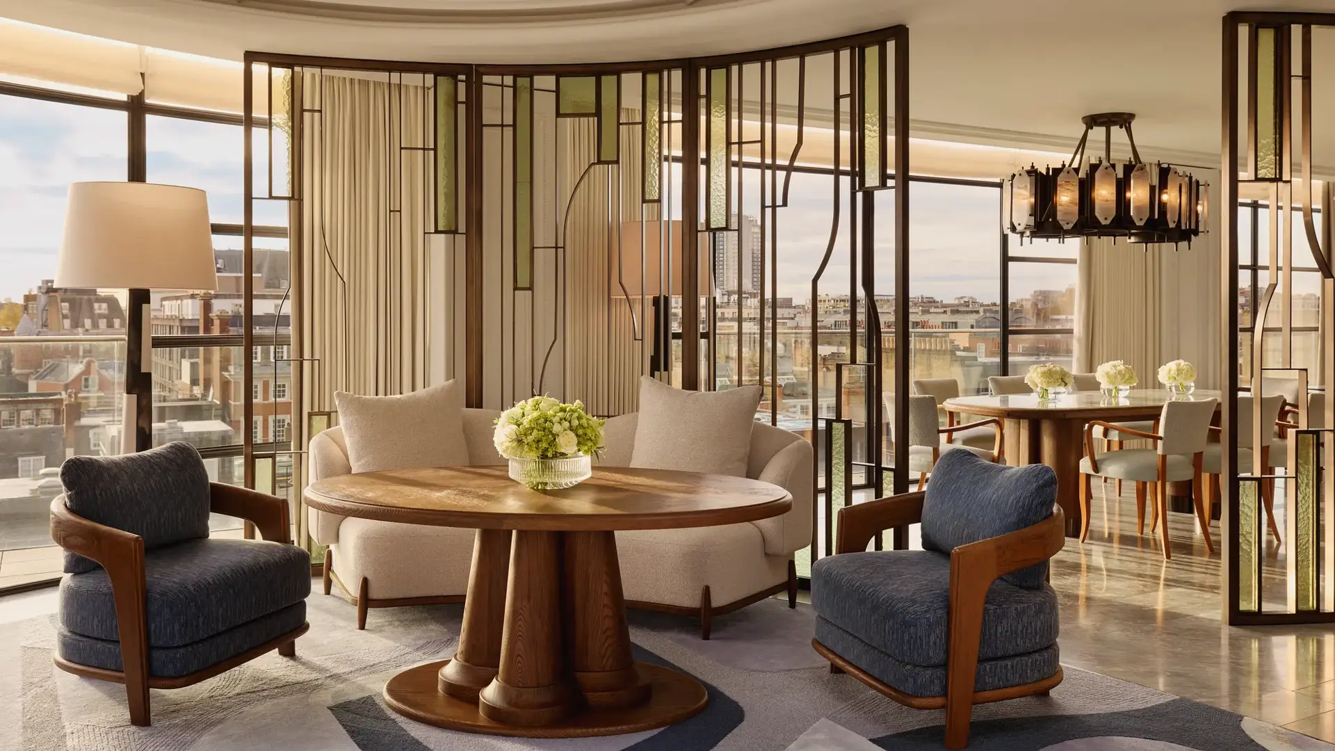 Bright open-plan living and dining area with large curved windows, featuring cream and blue armchairs around a round wooden table, decorative glass partitions, and a marble dining table beneath a statement chandelier.