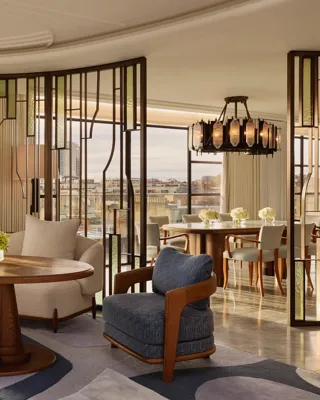 Bright open-plan living and dining area with large curved windows, featuring cream and blue armchairs around a round wooden table, decorative glass partitions, and a marble dining table beneath a statement chandelier.