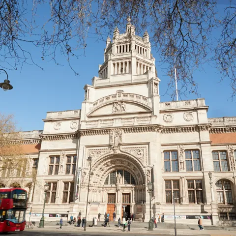 Victoria and Albert Museum facade in London framed by bare trees, with red double-decker buses passing in front.