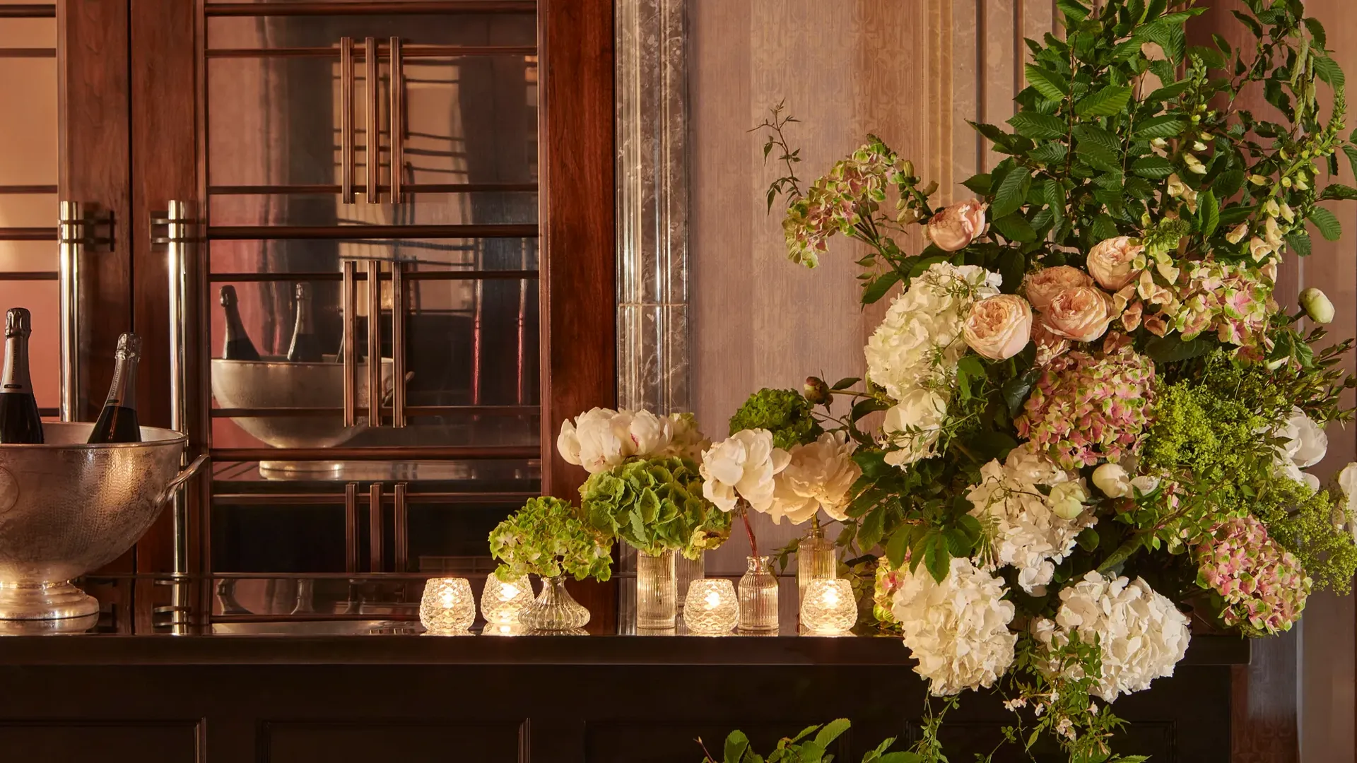 Elegant floral arrangement of hydrangeas and roses beside candlelit vases on a polished counter, with champagne bottles in a silver cooler behind.