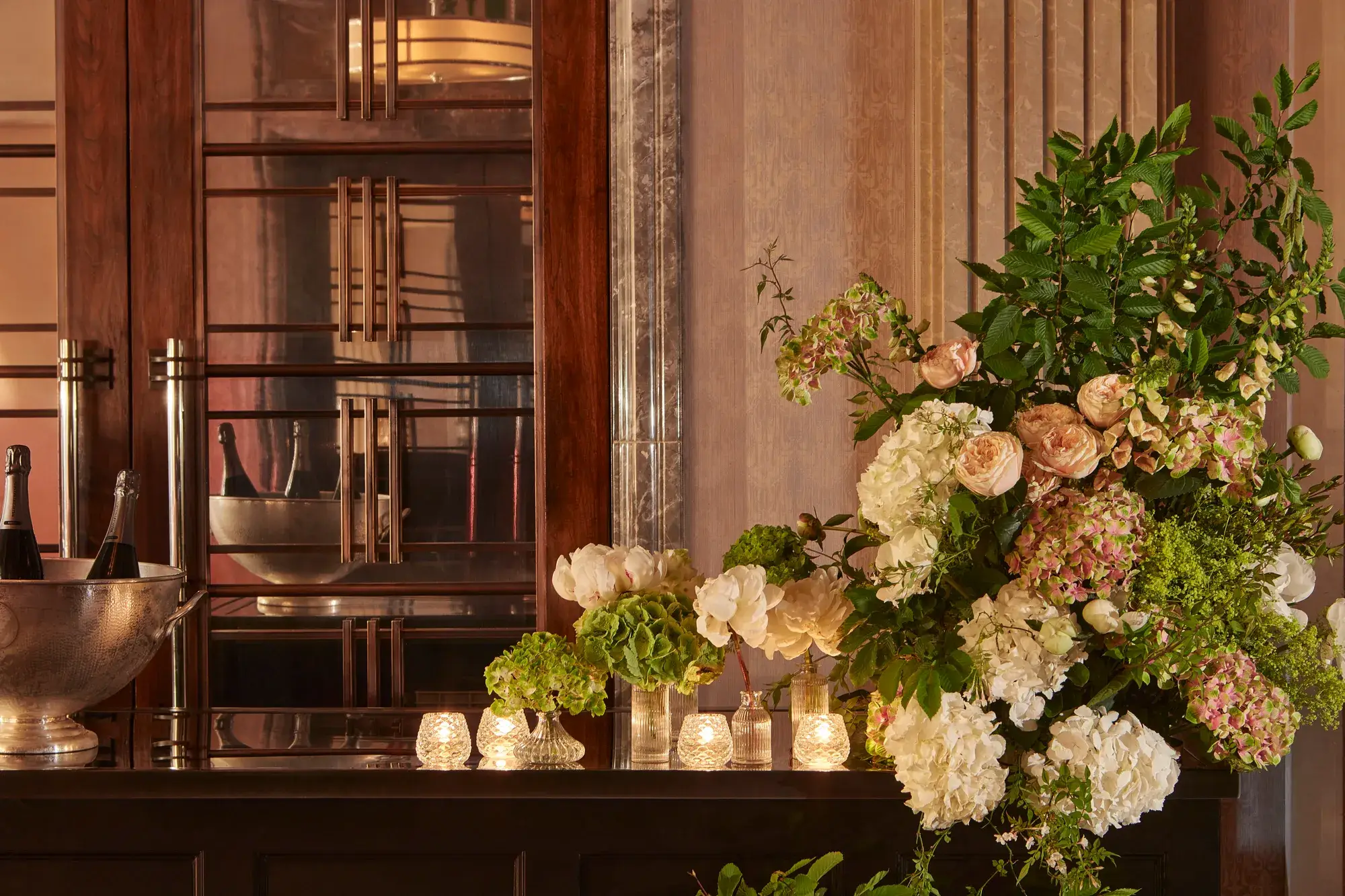 Elegant floral arrangement of hydrangeas and roses beside candlelit vases on a polished counter, with champagne bottles in a silver cooler behind.