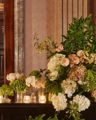 Elegant floral arrangement of hydrangeas and roses beside candlelit vases on a polished counter, with champagne bottles in a silver cooler behind.