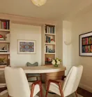 Home office with built-in bookshelves, a light wood desk, two cream chairs with wooden legs, and framed artwork on light walls.
