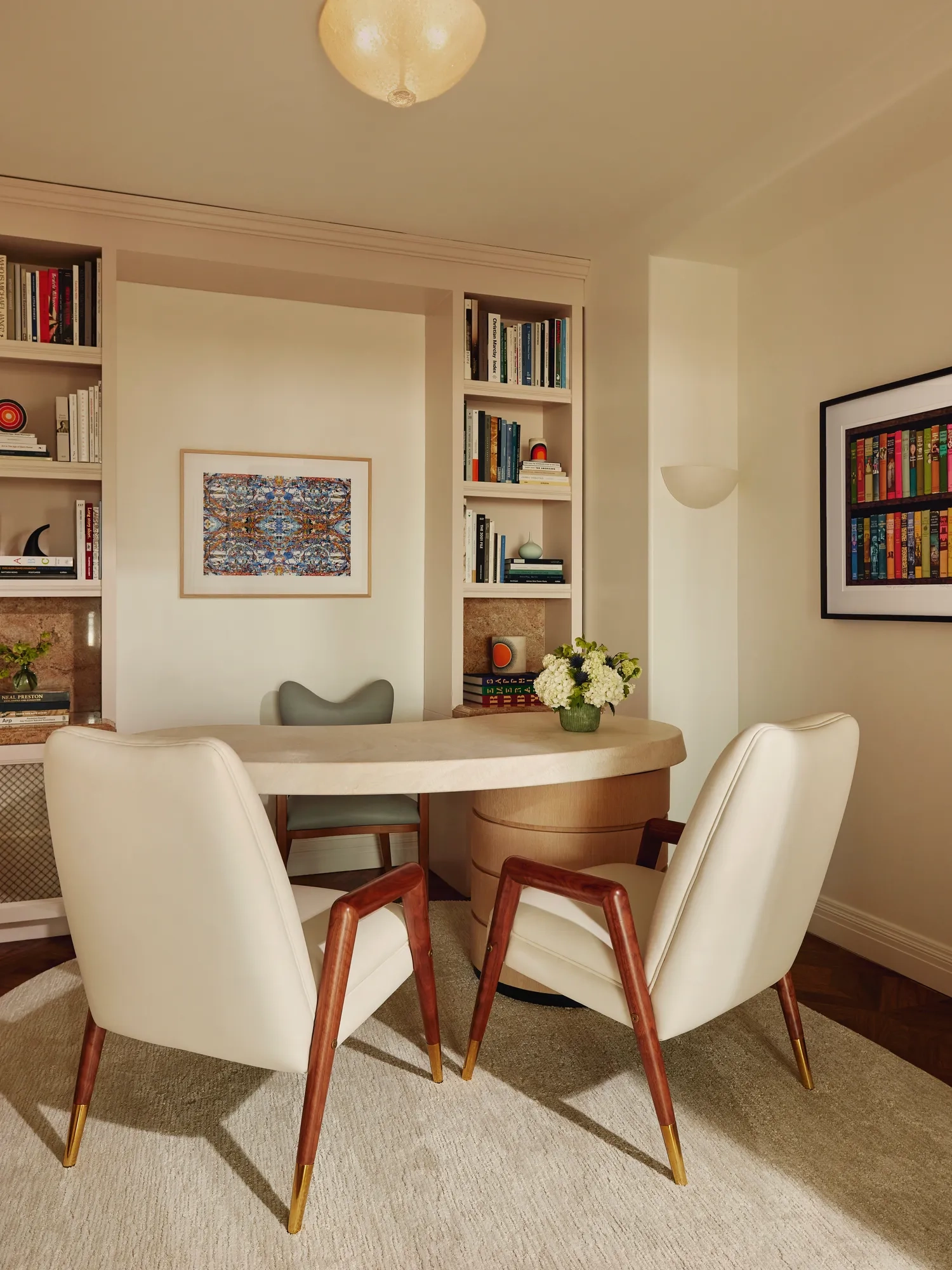 Home office with built-in bookshelves, a light wood desk, two cream chairs with wooden legs, and framed artwork on light walls.
