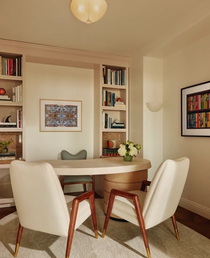 Home office with built-in bookshelves, a light wood desk, two cream chairs with wooden legs, and framed artwork on light walls.