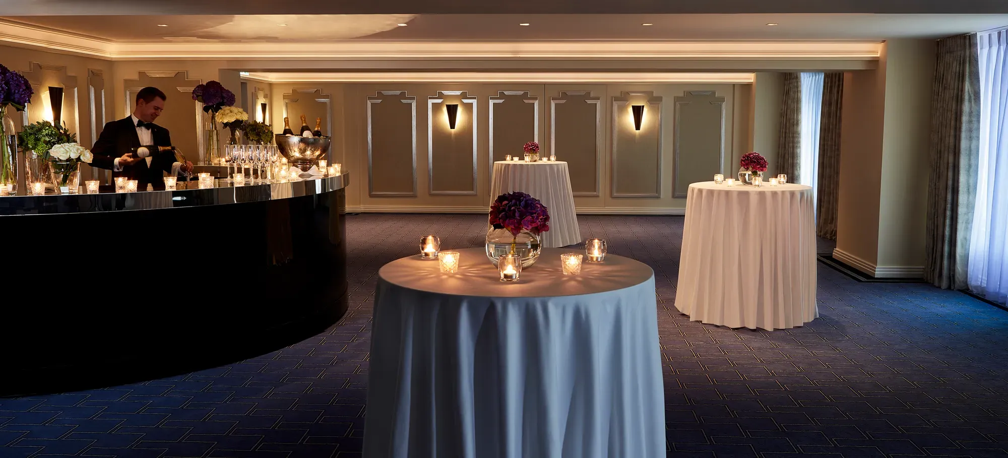 Intimate cocktail reception setup at Claridge’s with round candlelit tables draped in white cloths, floral centrepieces, and a bartender serving drinks from a curved black bar.