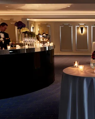 Intimate cocktail reception setup at Claridge’s with round candlelit tables draped in white cloths, floral centrepieces, and a bartender serving drinks from a curved black bar.