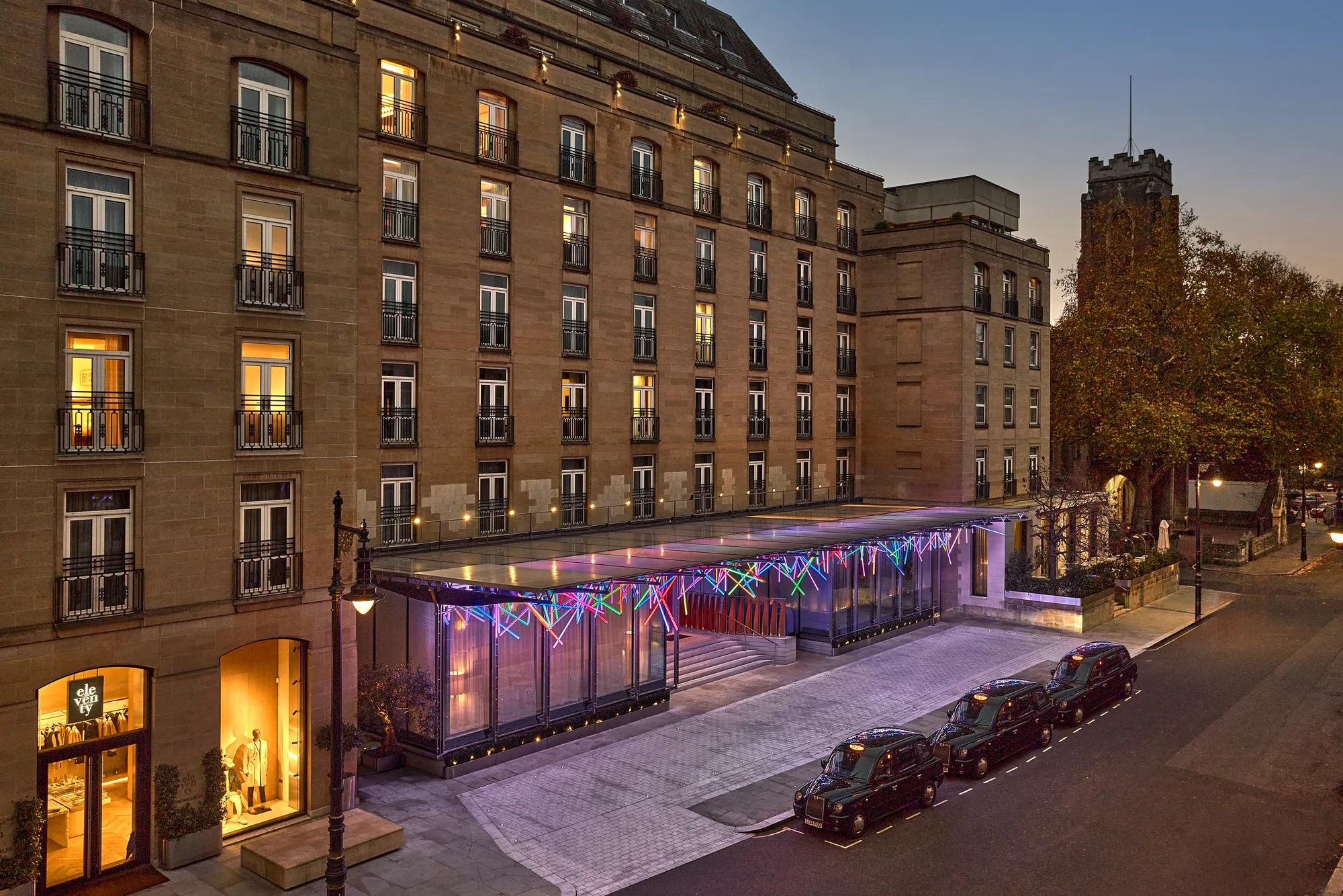 Hotel exterior at dusk with illuminated glass entrance canopy, parked taxis and lit windows along the street.