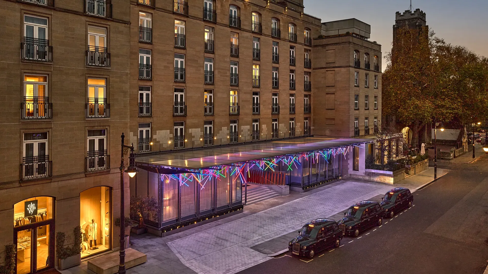 Hotel exterior at dusk with illuminated glass entrance canopy, parked taxis and lit windows along the street.