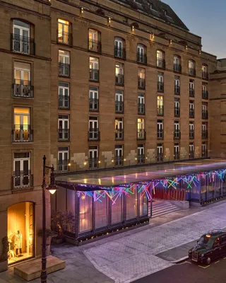 Hotel exterior at dusk with illuminated glass entrance canopy, parked taxis and lit windows along the street.