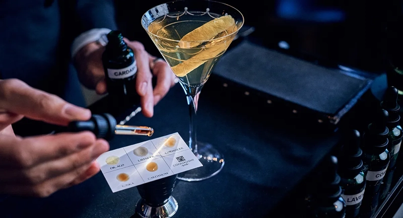 Bartender adding aromatic bitters with dropper beside martini glass garnished with citrus peel on bar counter.