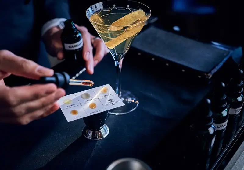 Bartender adding aromatic bitters with dropper beside martini glass garnished with citrus peel on bar counter.