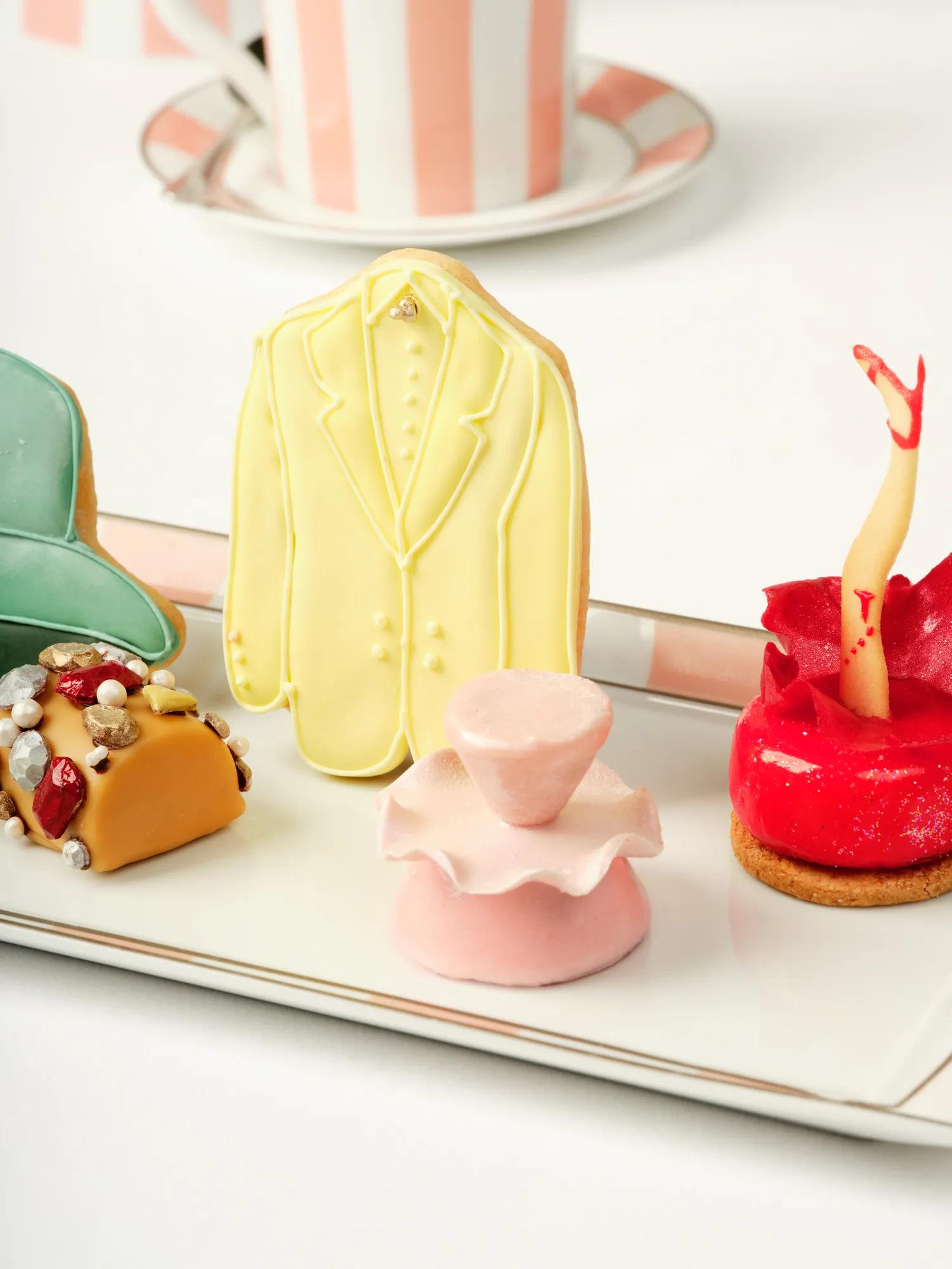 Assorted fashion‑inspired pastries on a tray, including a yellow suit cookie, pink ruffled treat, and red dessert with decorative leg topper.