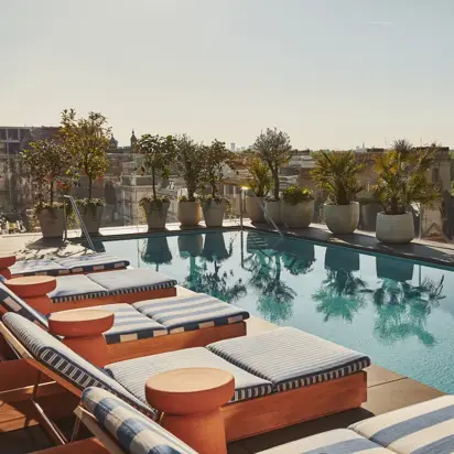 Rooftop pool lined with blue-striped loungers and potted plants, overlooking the city skyline and treetops.