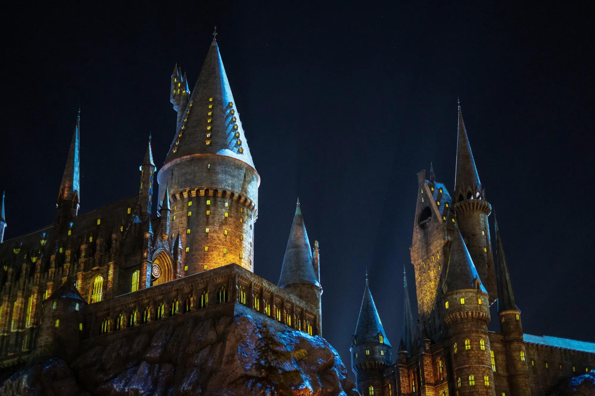 Illuminated castle with soaring towers and glowing windows at night, set against a dark sky and rocky landscape.