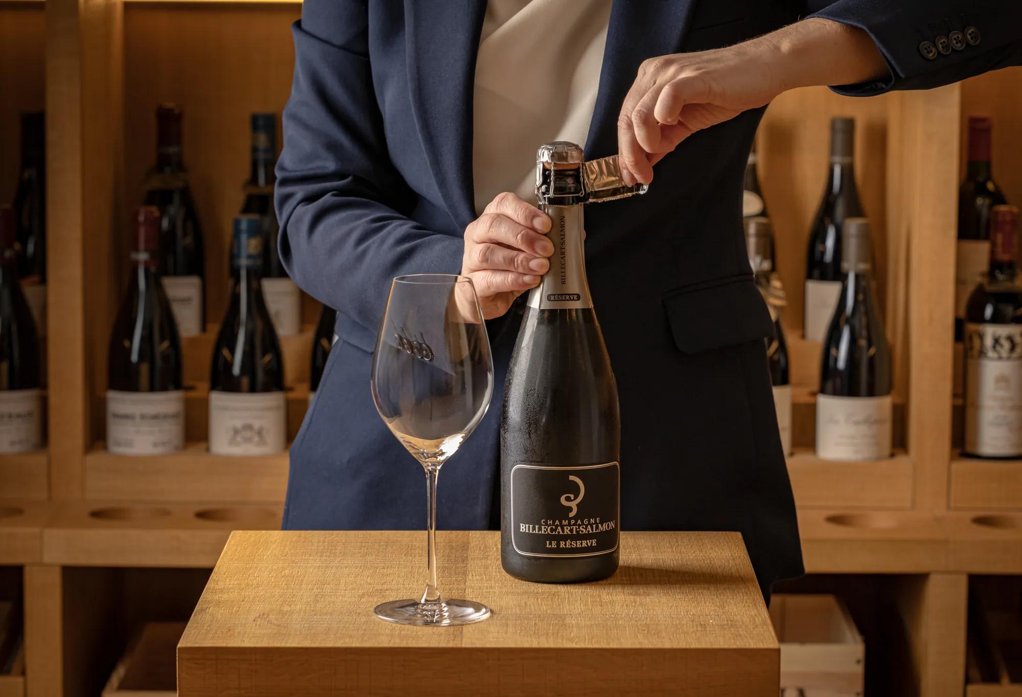 A bottle of Champagne is opened beside an empty wine glass, with rows of wine bottles displayed on wooden shelves in the background.