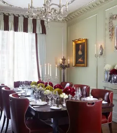 Elegant private dining room with a dark wood table, red leather chairs, floral centrepieces and classic portraits, lit by a chandelier.