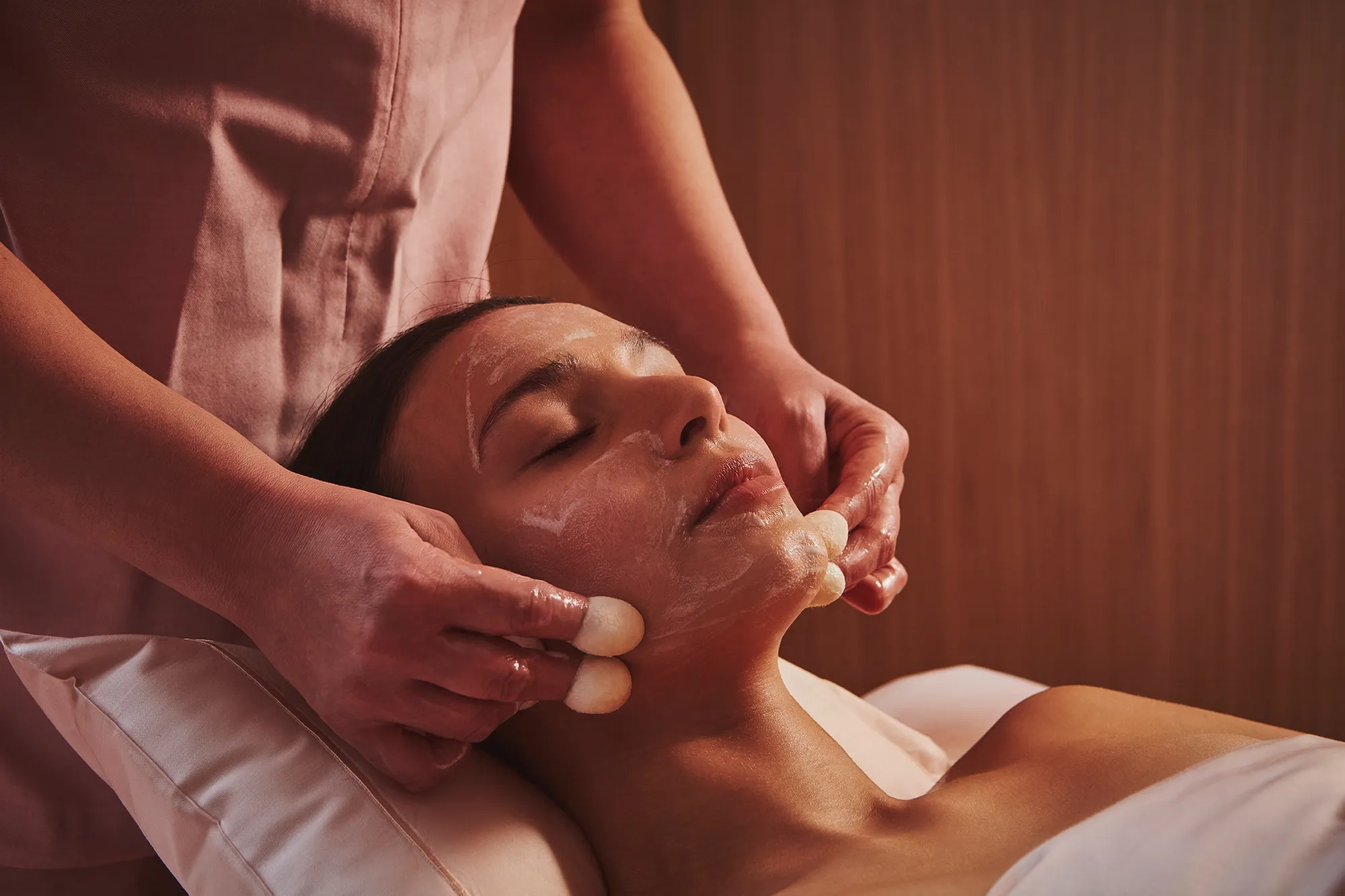 A woman receives a soothing facial treatment with a creamy mask and massage tools from a therapist in a softly lit spa room at The Maybourne Riviera.