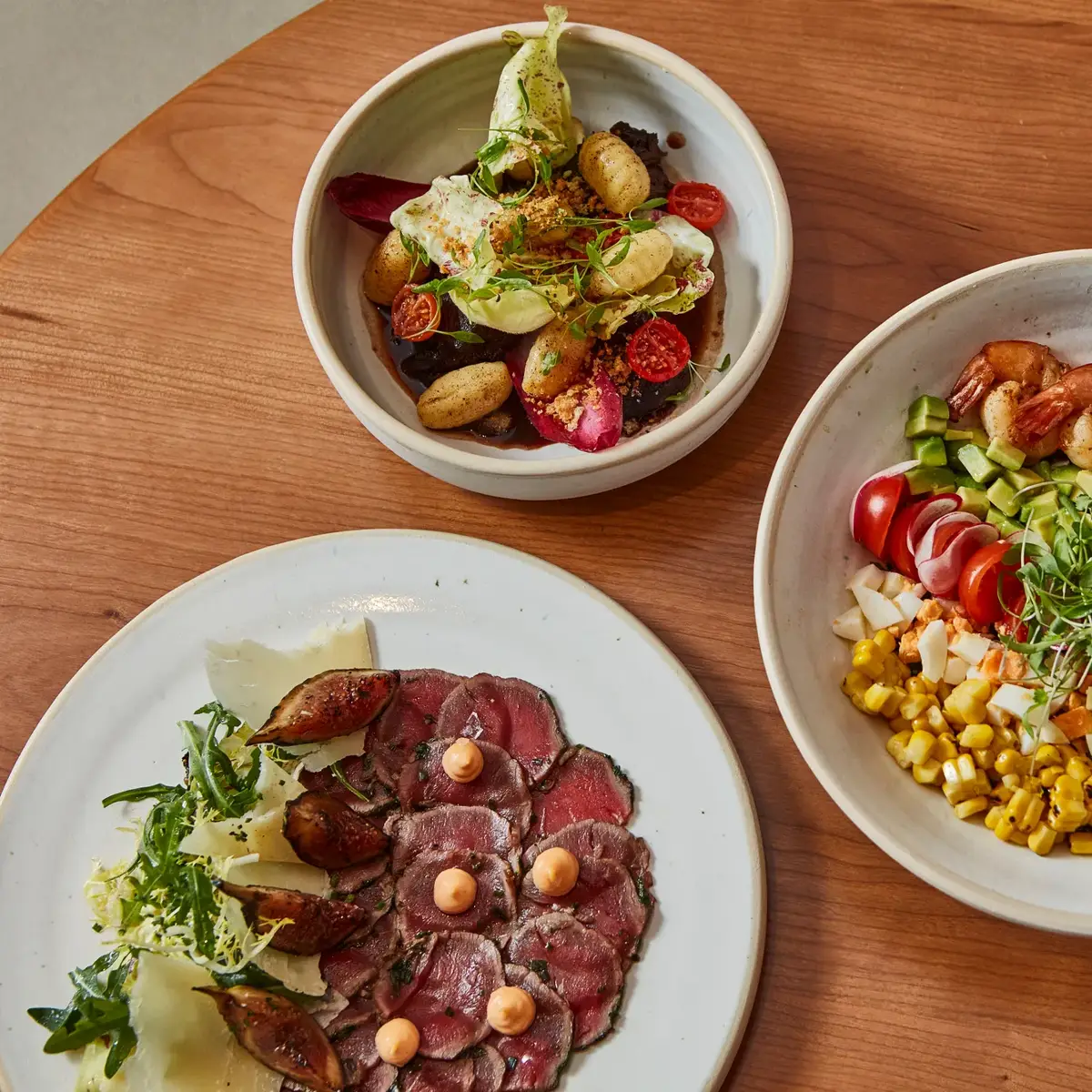 Four plated dishes on a wooden table, including shrimp salad, sliced beef with figs, and colourful vegetable plates.