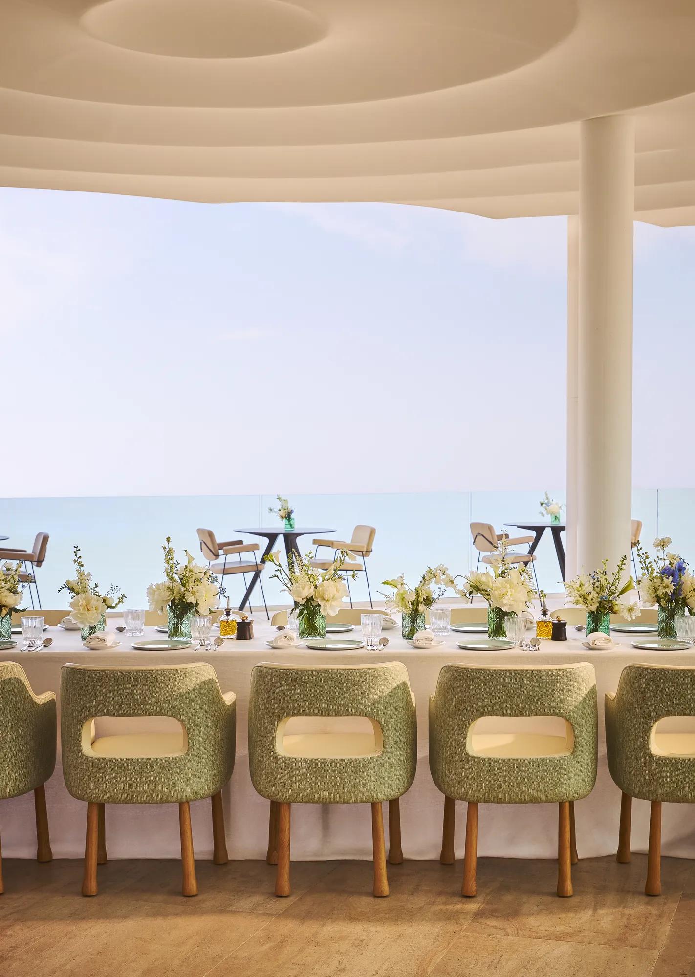 Salle à manger élégante avec des chaises vertes rembourrées, une nappe blanche et des vases de fleurs fraîches, face à de grandes baies vitrées offrant une vue sur la mer et une terrasse extérieure.