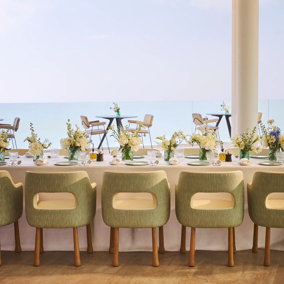 Salle à manger élégante avec des chaises vertes rembourrées, une nappe blanche et des vases de fleurs fraîches, face à de grandes baies vitrées offrant une vue sur la mer et une terrasse extérieure.
