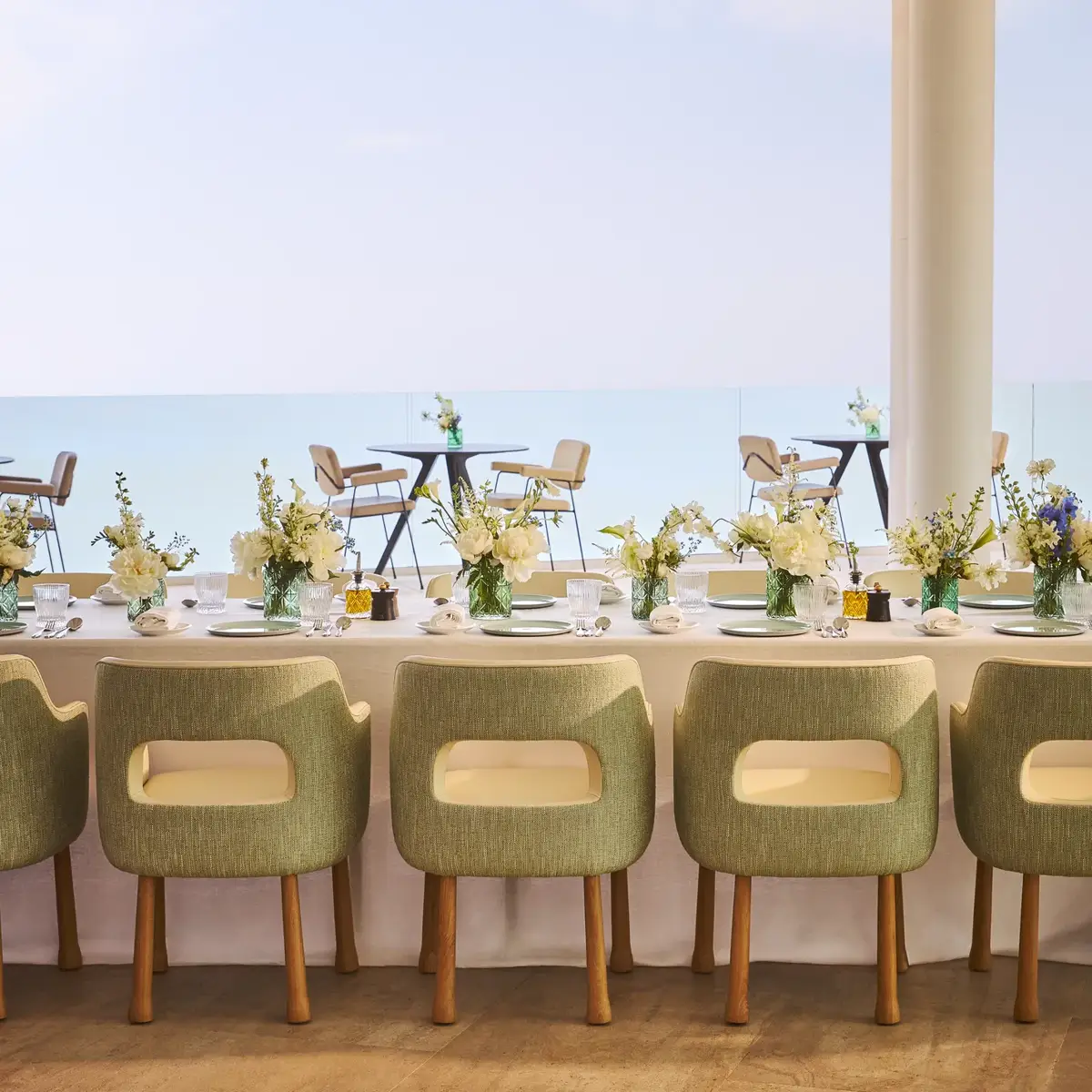 Salle à manger élégante avec des chaises vertes rembourrées, une nappe blanche et des vases de fleurs fraîches, face à de grandes baies vitrées offrant une vue sur la mer et une terrasse extérieure.
