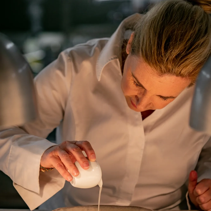 Chef pouring sauce over plated dish at marble counter beneath heat lamps in professional kitchen.