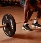 Person performing a barbell deadlift in a gym, with weight plates and dumbbells visible in the background.