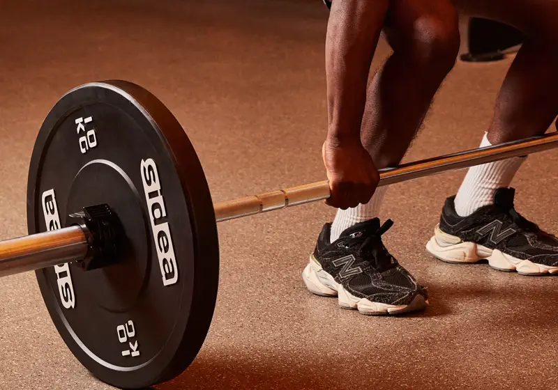 Person performing a barbell deadlift in a gym, with weight plates and dumbbells visible in the background.