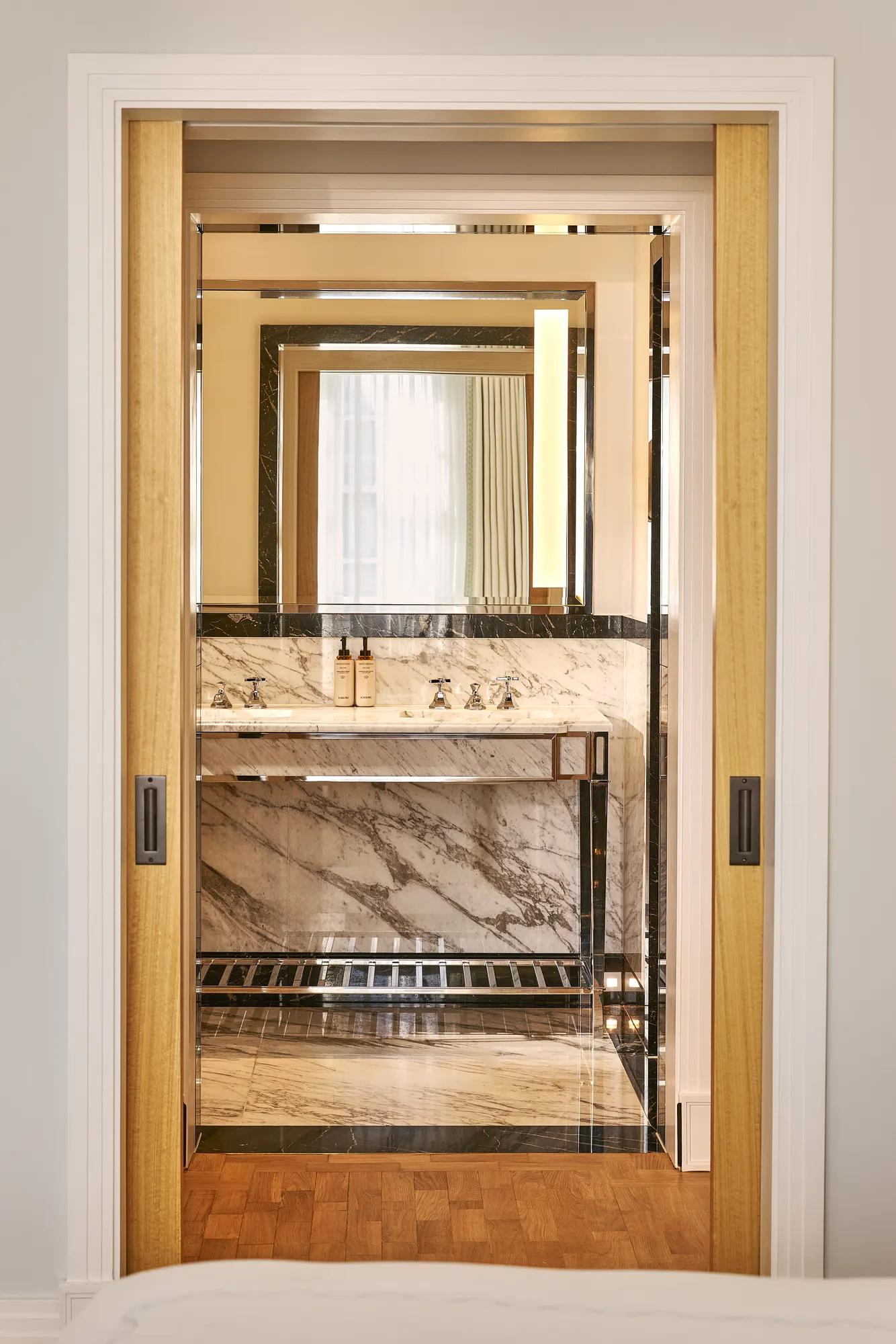 View through sliding doors into marble bathroom with vanity sink and illuminated mirror.
