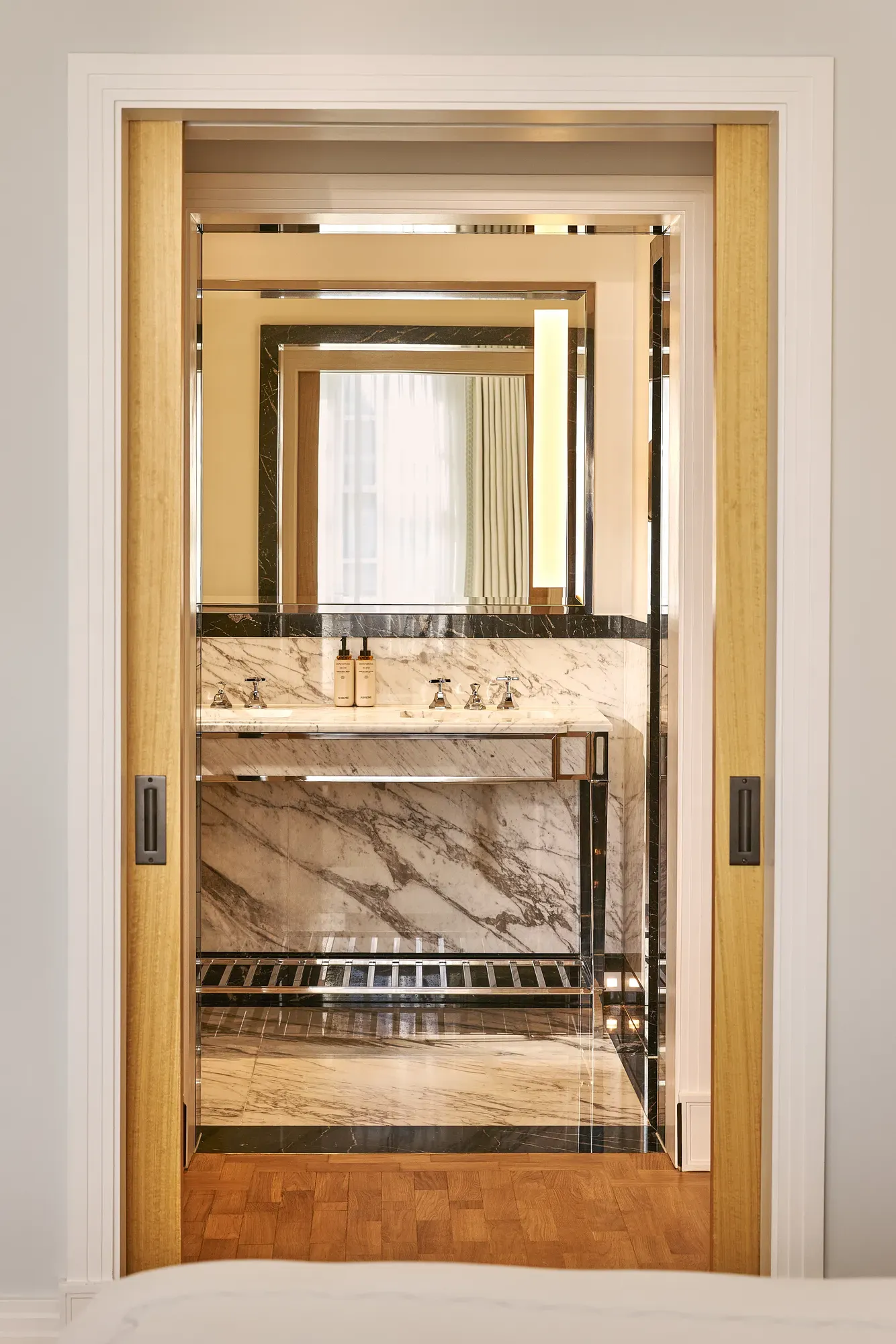 View through sliding doors into marble bathroom with vanity sink and illuminated mirror.