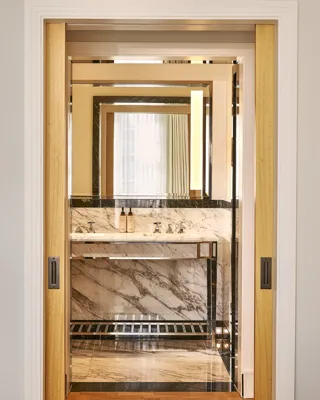 View through sliding doors into marble bathroom with vanity sink and illuminated mirror.