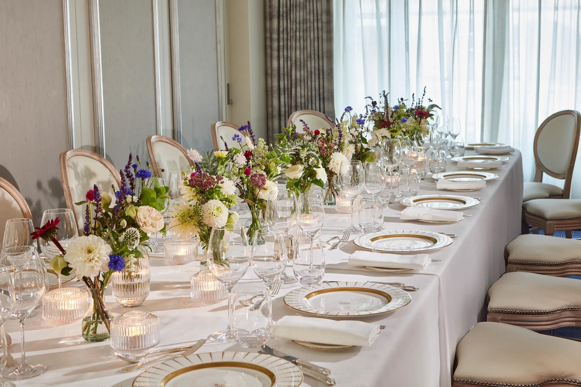 Elegant dining table at Claridge’s set with white linen, fine china, glassware, and floral centrepieces of white, purple, and pink blooms, illuminated by soft candlelight.