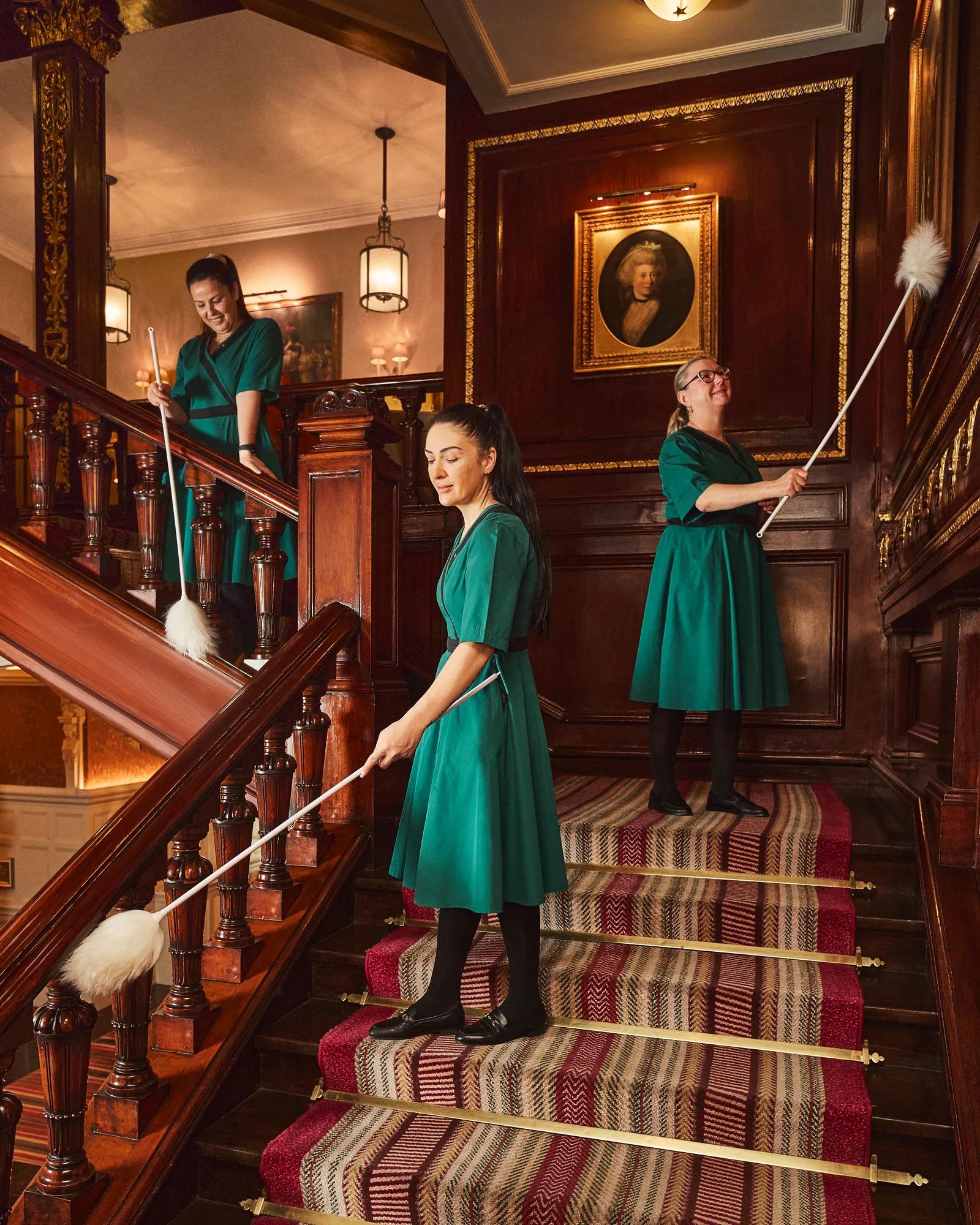 Three housekeeping staff dusting a polished wooden staircase in an elegant interior with dark wood panelling and framed artwork.
