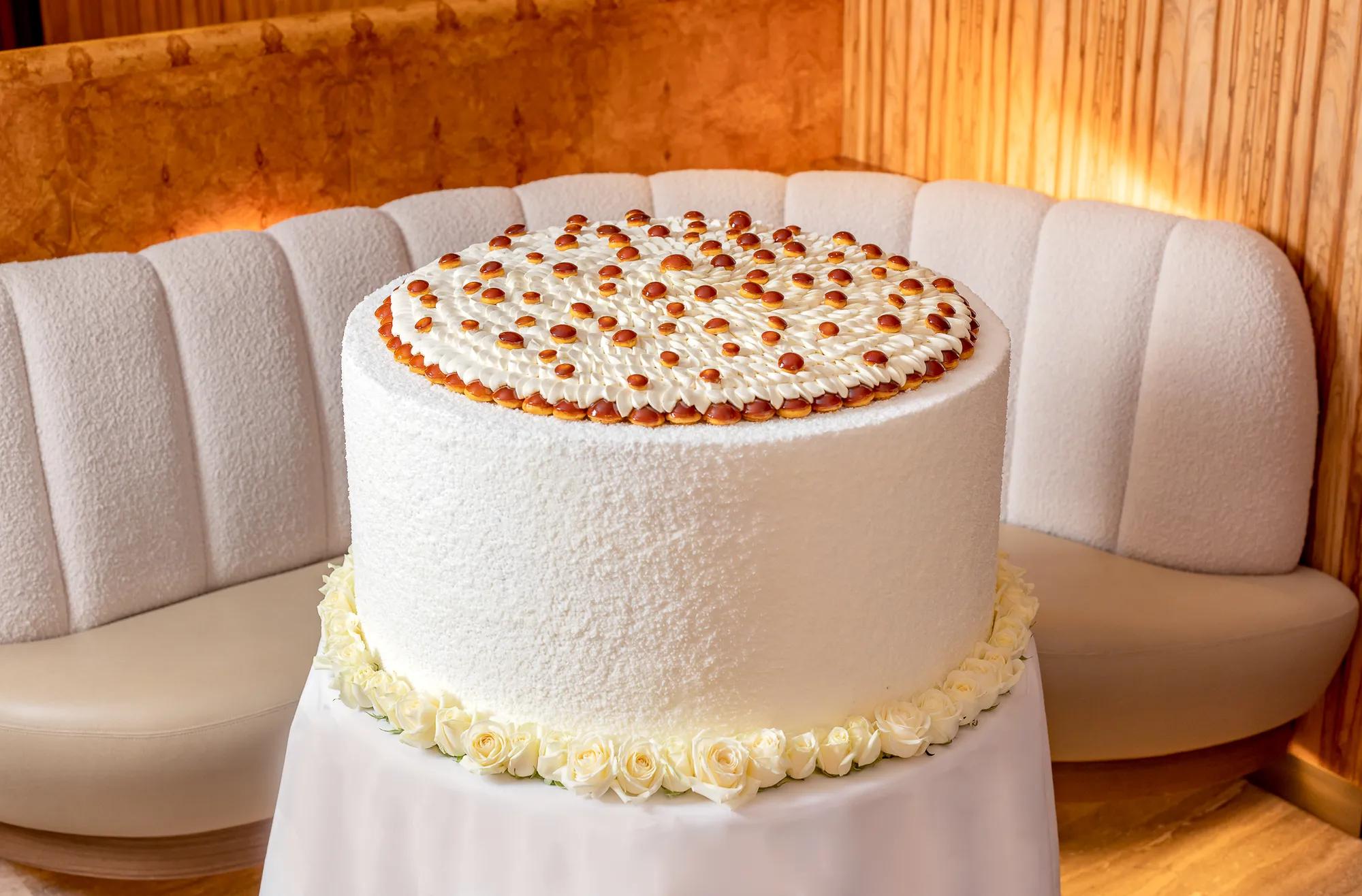 Large white frosted cake topped with piped cream and caramel drops, displayed on a table in a softly lit lounge.