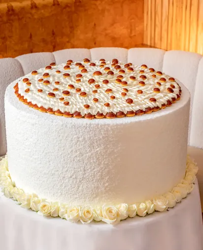 Large white frosted cake topped with piped cream and caramel drops, displayed on a table in a softly lit lounge.